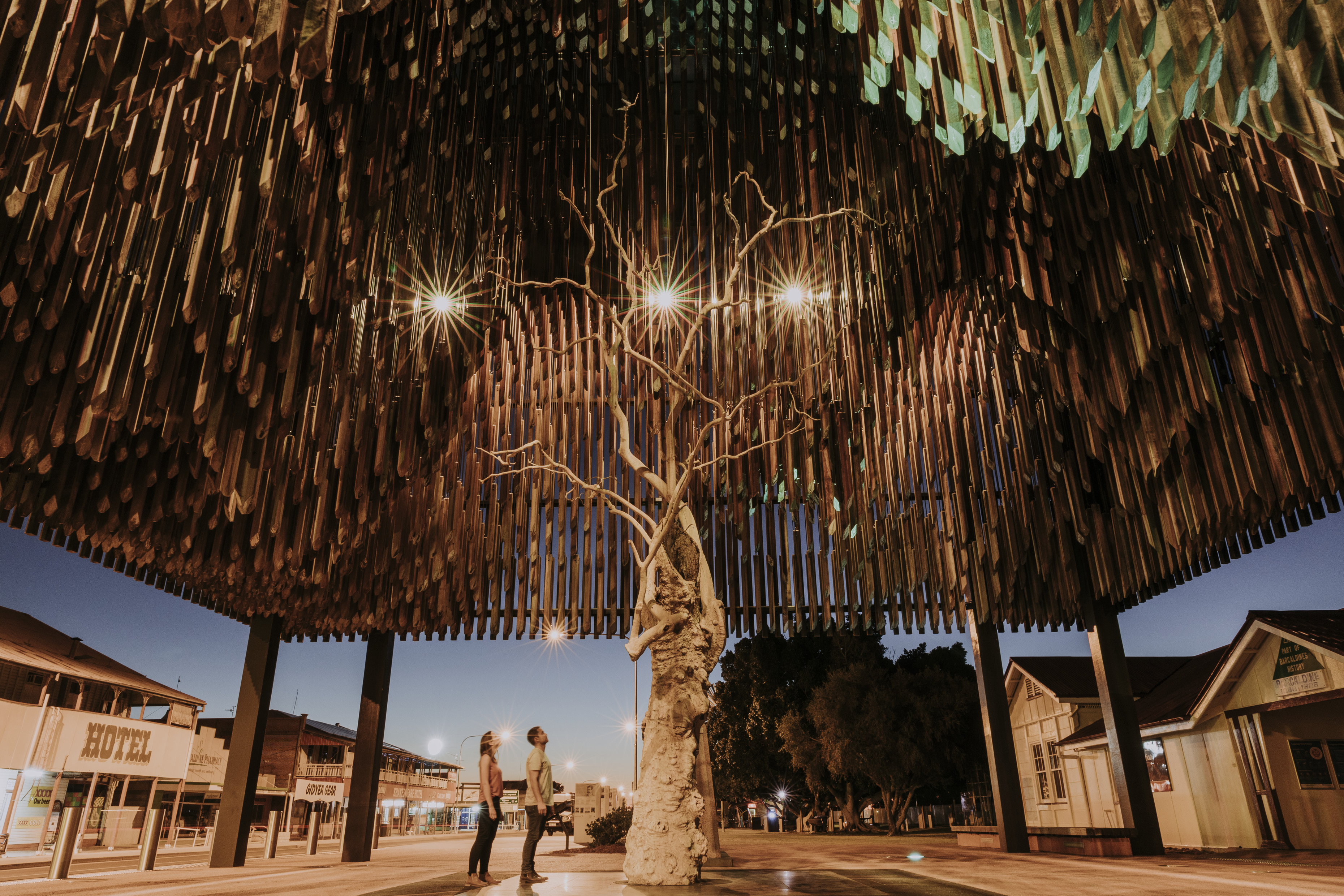 Two travellers are looking up at the Tree of Knowledge, a heritage listed ghost gum tree sits underneath a huge architecturally designed memorial